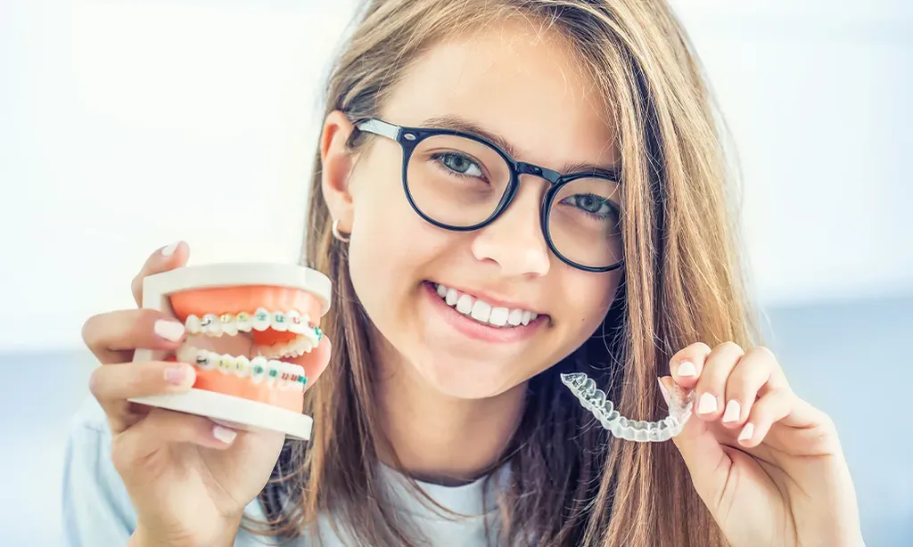 a smiling girl holding a model of a mouth with braces in the left hand and a clear appliance in the right hand.